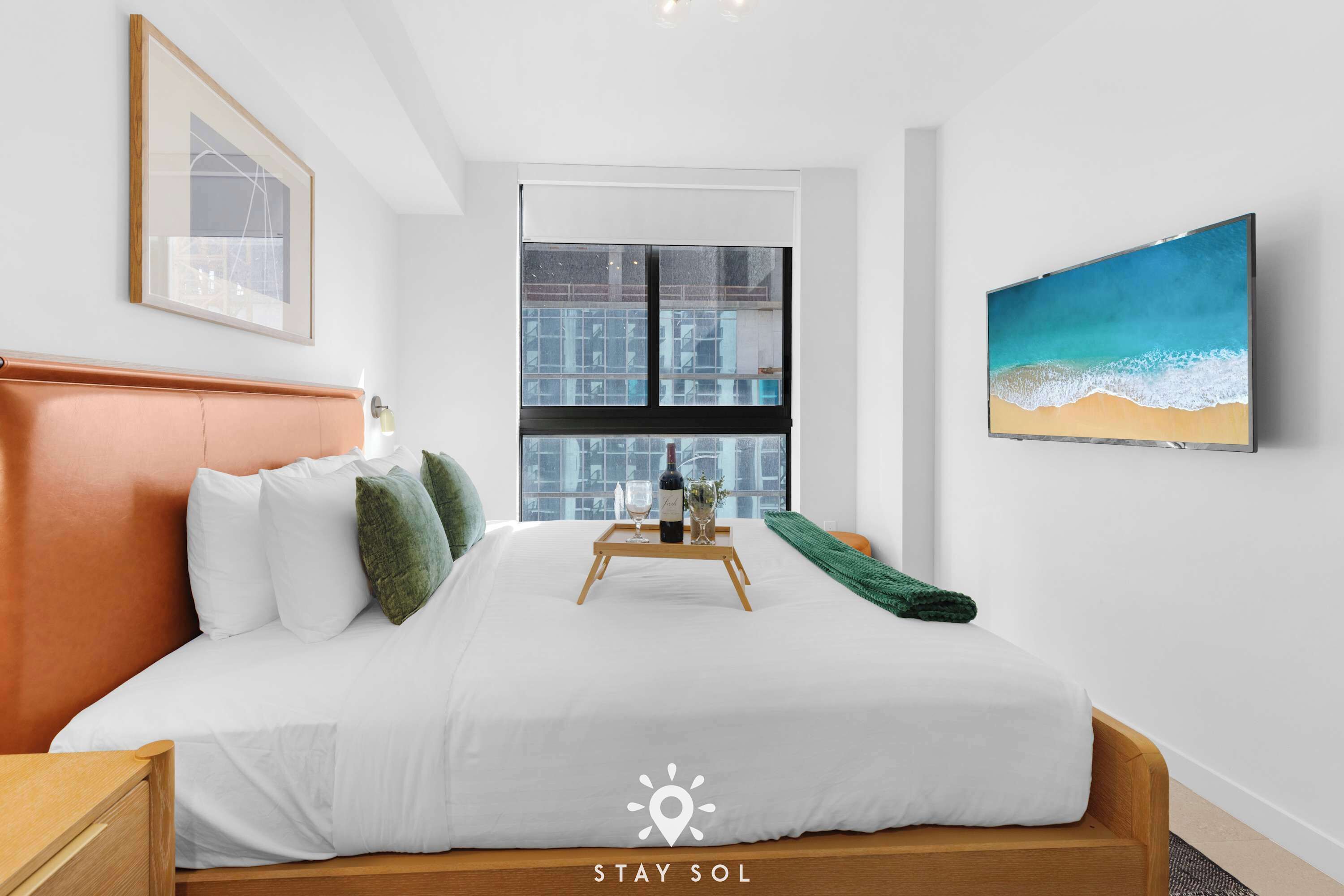 A hotel room with a bed, a window with a city view, a painting of a beach, and a tray with glasses and a bottle.