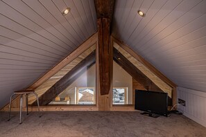 A sloped ceiling attic room with a television, a stool, and a window with a view of a house. - Toits des Pècles – Stylish Alpine Loft with Mont Blanc Views (Chamonix-Mont-Blanc)