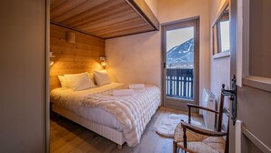 A cozy bedroom with a wooden ceiling, a large bed with white bedding, a wooden chair, and a view of mountains through a window. - Balcons de Champraz- Spectacular Mont Blanc and Lake Views (Chamonix-Mont-Blanc)