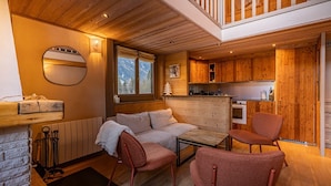A cozy cabin interior with a sofa, two chairs, a small table, a kitchen area, and a window with a view of the mountains. - Balcons de Champraz- Spectacular Mont Blanc and Lake Views (Chamonix-Mont-Blanc)