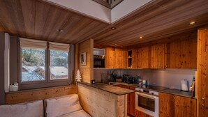 A cozy cabin interior with a wooden interior, a sofa, a kitchen with wooden cabinets, and a window with a snowy view. - Balcons de Champraz- Spectacular Mont Blanc and Lake Views (Chamonix-Mont-Blanc)