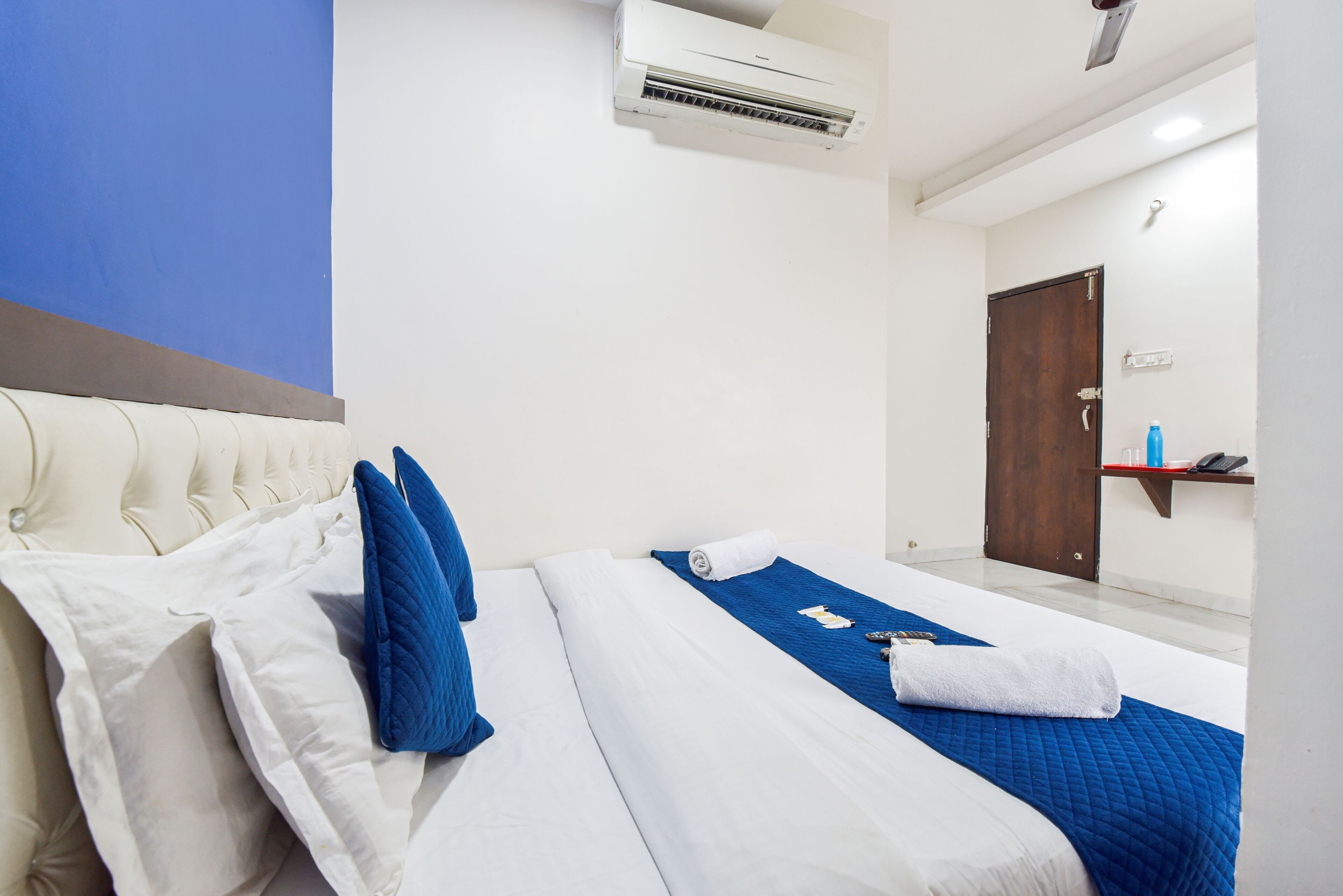 Une chambre d’hôtel avec un lit, des oreillers, une couverture bleue, une serviette blanche, un climatiseur, une porte en bois et une petite étagère sur laquelle se trouvent une bouteille et un téléphone.