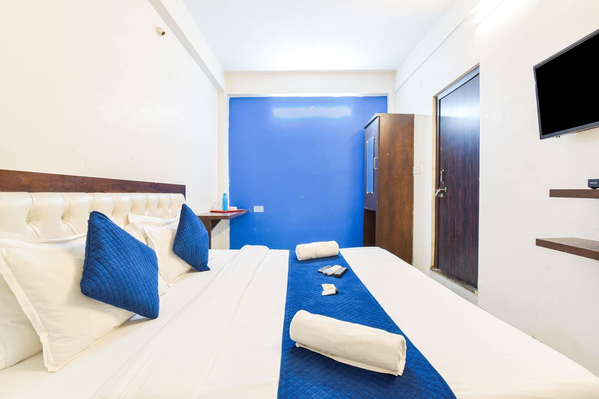 A hotel room with a large bed, a blue accent wall, a wooden wardrobe, and a flat-screen TV mounted on the wall.