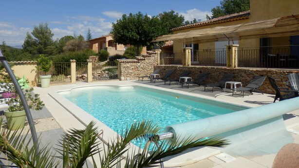 A pool area with a stone wall, lounge chairs, and a yellow building with a tiled roof. - Gîte des Lavandes (MONTAGNAC MONTPEZAT)