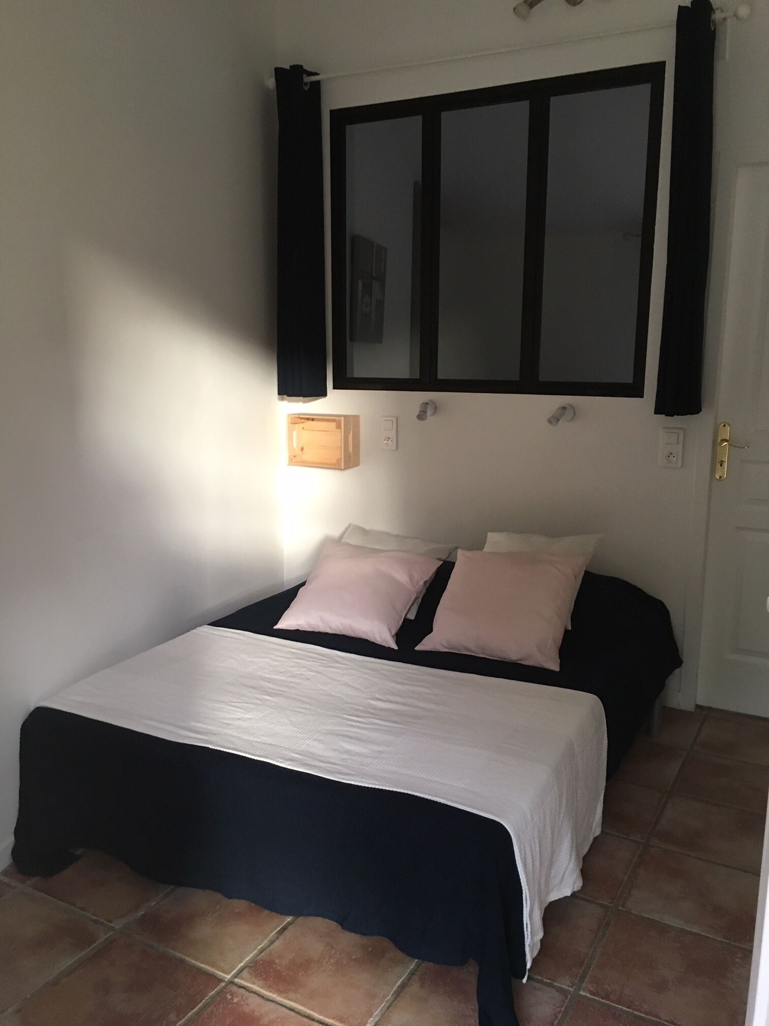 A bedroom with a bed, two bedside tables, a window with curtains, and a wall-mounted light.
