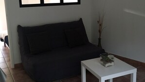 A living room with a dark-colored sofa, a white coffee table, and a large window with blinds. - Gîte des Lavandes (MONTAGNAC MONTPEZAT)