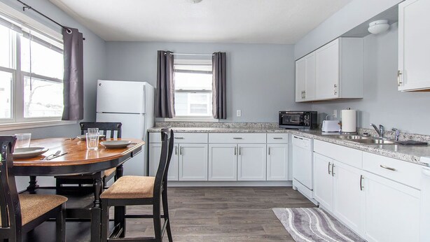 A kitchen with white cabinets, a dining table with chairs, a microwave, and a refrigerator. - 2BR I 4 mins to FargoDome, Free Pking, Queen x 2 (Fargo)