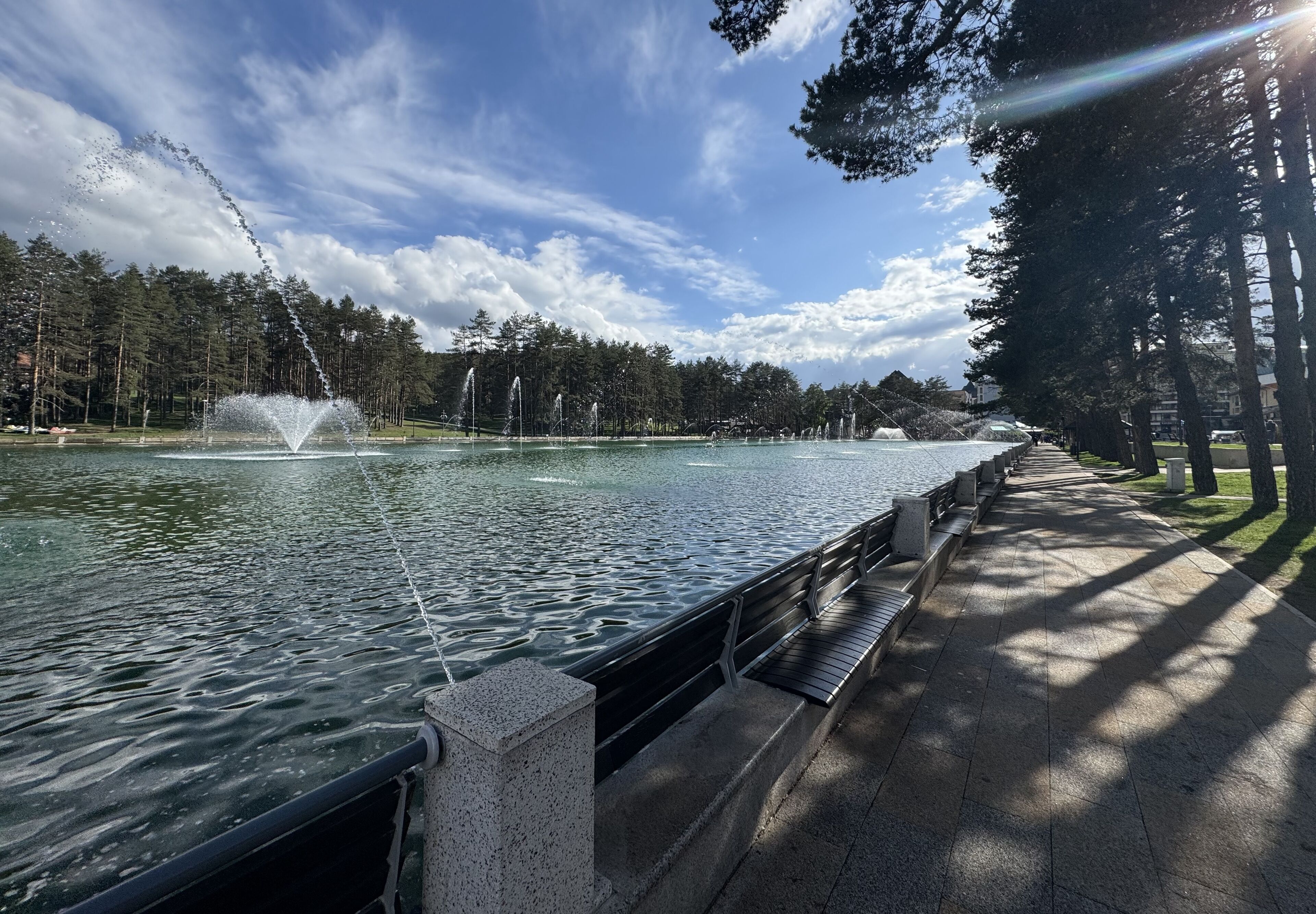 A serene lakeside walkway with a fountain, surrounded by trees and a clear sky.