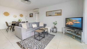 A modern living room with a grey sofa, a coffee table, a TV on a stand, a patterned rug, and a dining area with chairs and a table. - Le Panama (Saint-Raphaël)