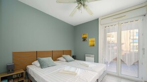 A bedroom with two beds, a wooden headboard, a nightstand, a ceiling fan, and a sliding glass door to a balcony. - Le Panama (Saint-Raphaël)