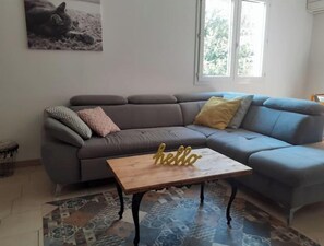 Un salon avec un canapé gris, une table basse en bois décorée du mot «
bonjour
», un tapis à motifs et un tableau représentant un chat accroché au mur.