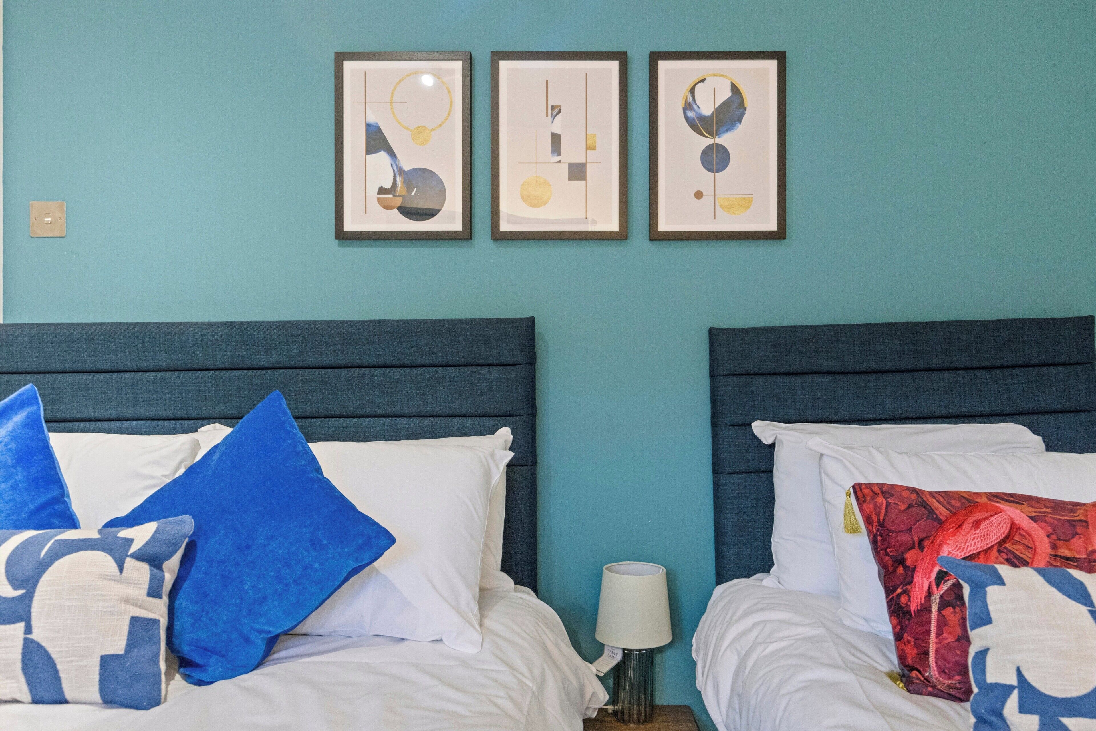 A hotel room with two beds, each with a headboard and pillows. There are three framed artworks on the wall above the beds.