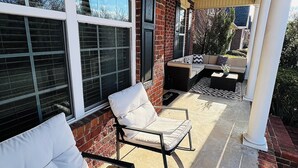 Ein überdachter Patio-Bereich mit zwei weißen, gepolsterten Stühlen, einem kleinen Tisch und einer Ziegelwand.