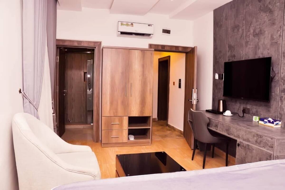 Moderni hotellihuone, jossa on puinen vaatekaappi, työpöytä tuolilla, seinälle asennettu litteänäyttöinen televisio ja ilmastointilaite.