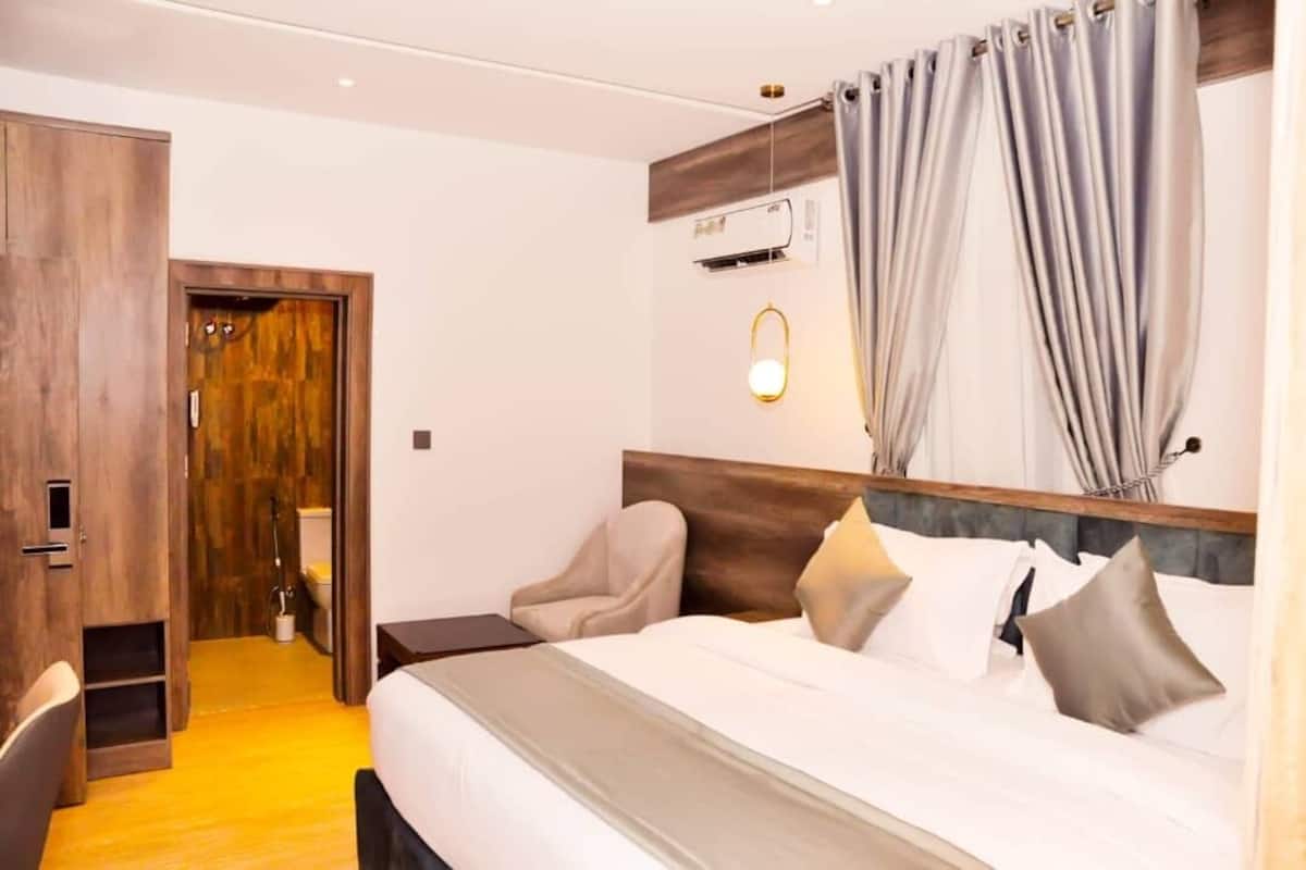 Une chambre d’hôtel avec une armoire en bois, un lit avec une literie blanche, une tête de lit, une table de chevet et un climatiseur fixé au mur.