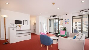 Camera d'albergo moderna con tappeto rosso, reception, poltrona blu, divano beige e un'ampia finestra con vista sulla città.