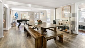 Un comedor amplio con una larga mesa de madera y bancos, una pared blanca con espejo, y vista al exterior a través de una ventana.