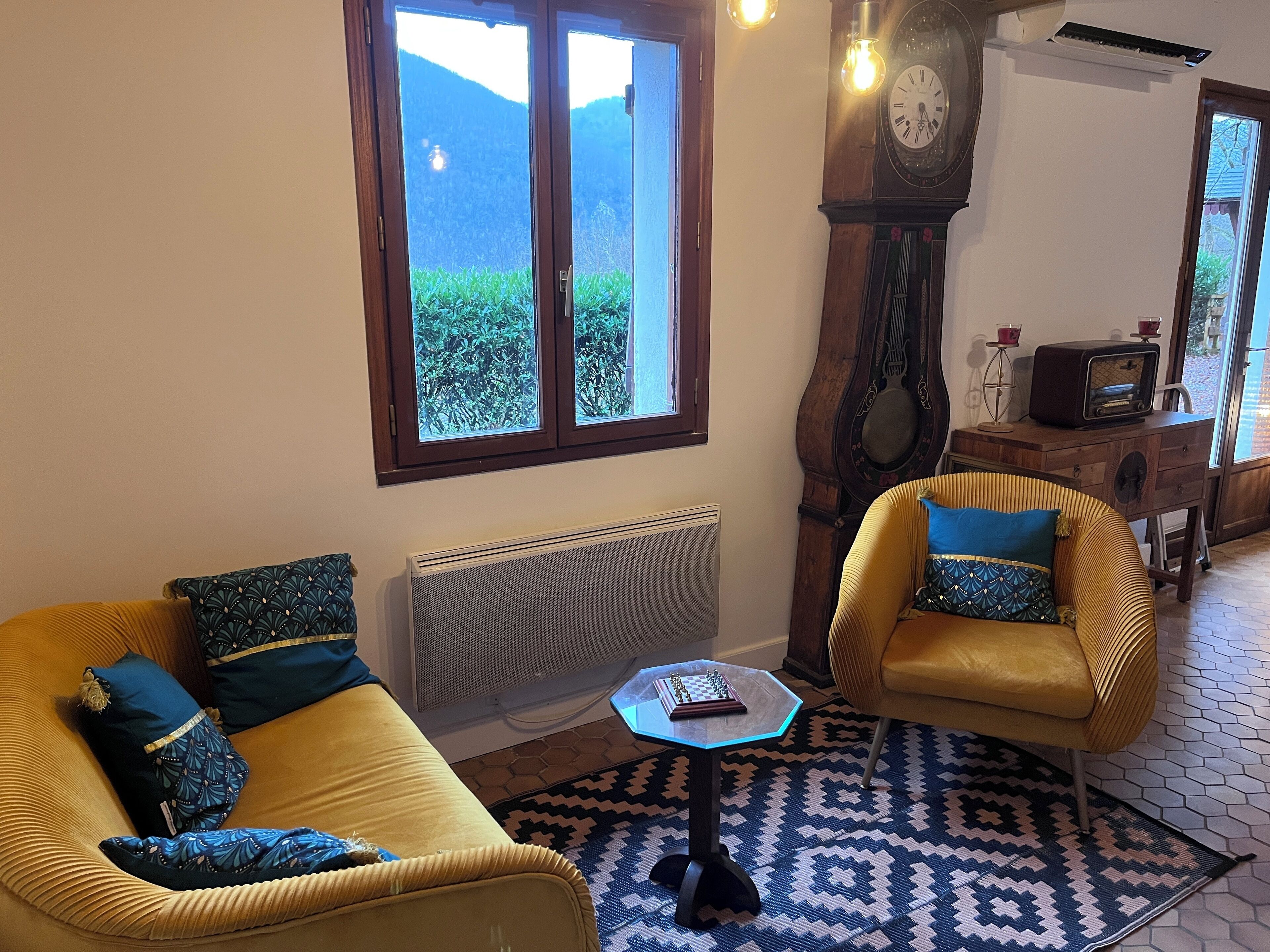 Una sala de estar acogedora con un sofá, un sillón y una mesita. Un reloj grande y una radio antigua están sobre un aparador de madera.
