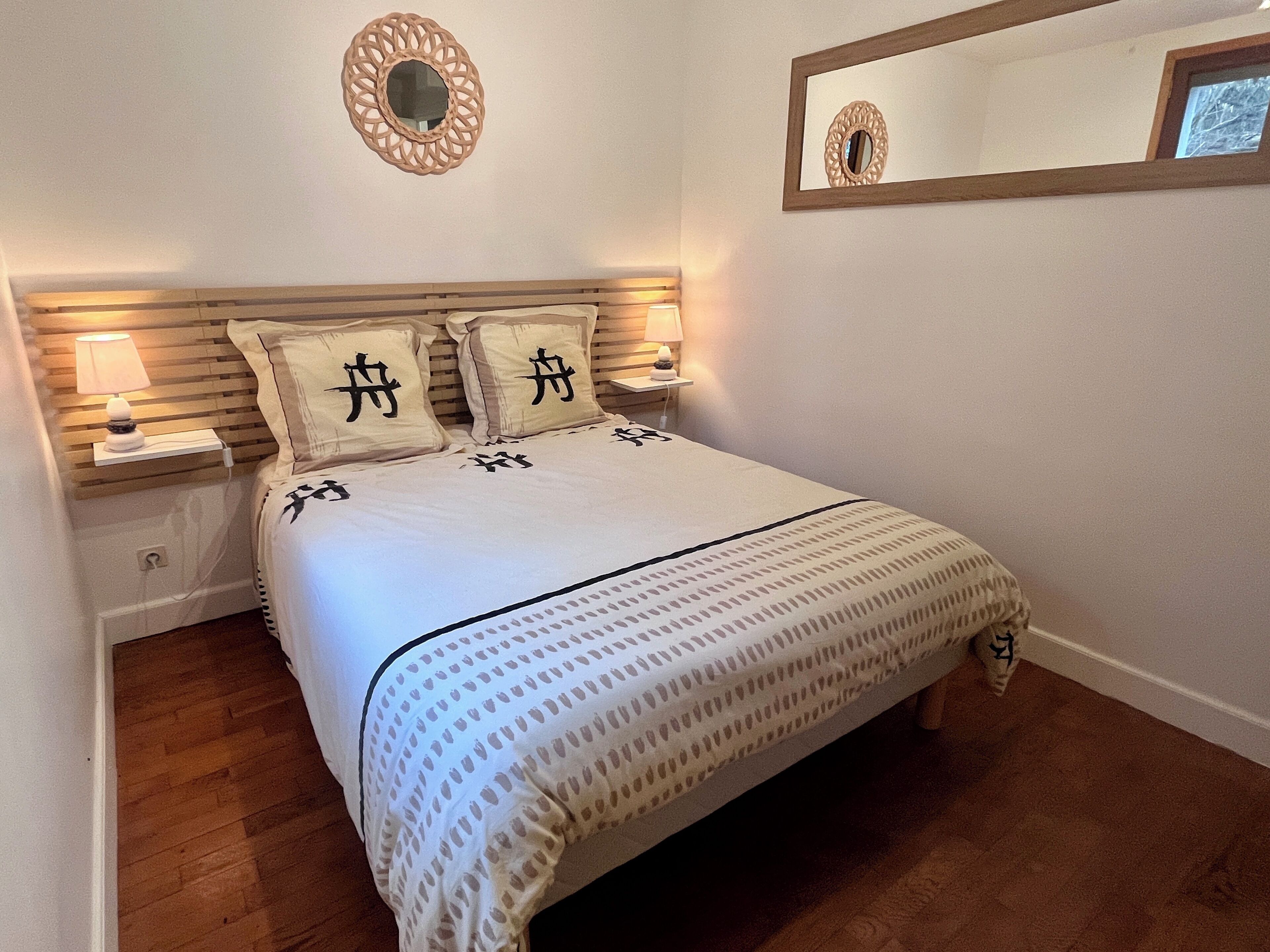 Un lit bien fait, avec une couvre-lit à motifs et des oreillers ornés de caractères chinois, une lampe de chevet et un miroir fixé au mur.