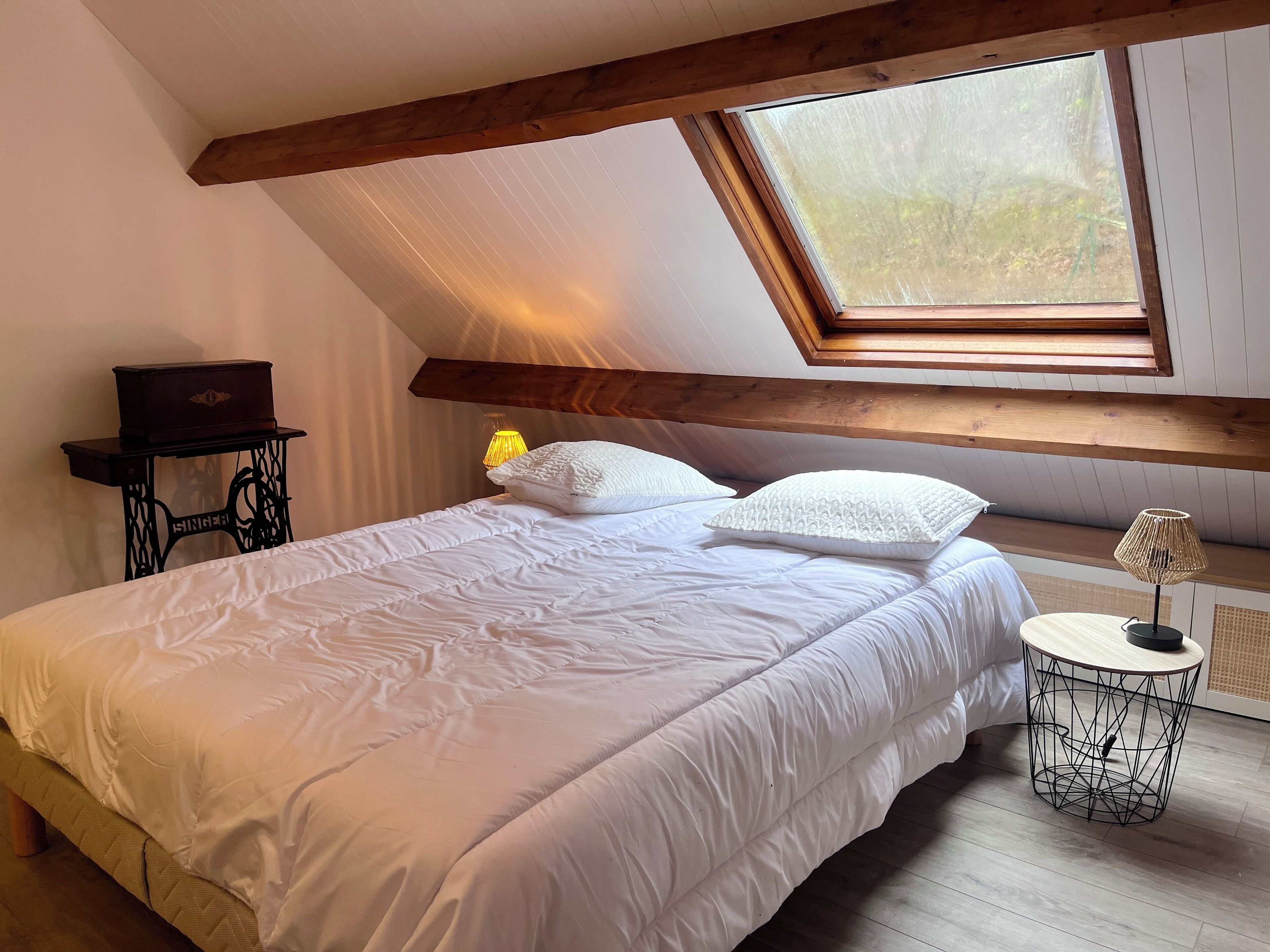 Una camera da letto con soffitto spiovente, un lucernario, un letto con biancheria bianca, un tavolino con lampada e un armadietto in legno.