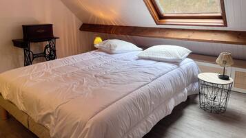 Una camera da letto con soffitto spiovente, un lucernario, un letto con biancheria bianca, un tavolino con lampada e un armadietto in legno.