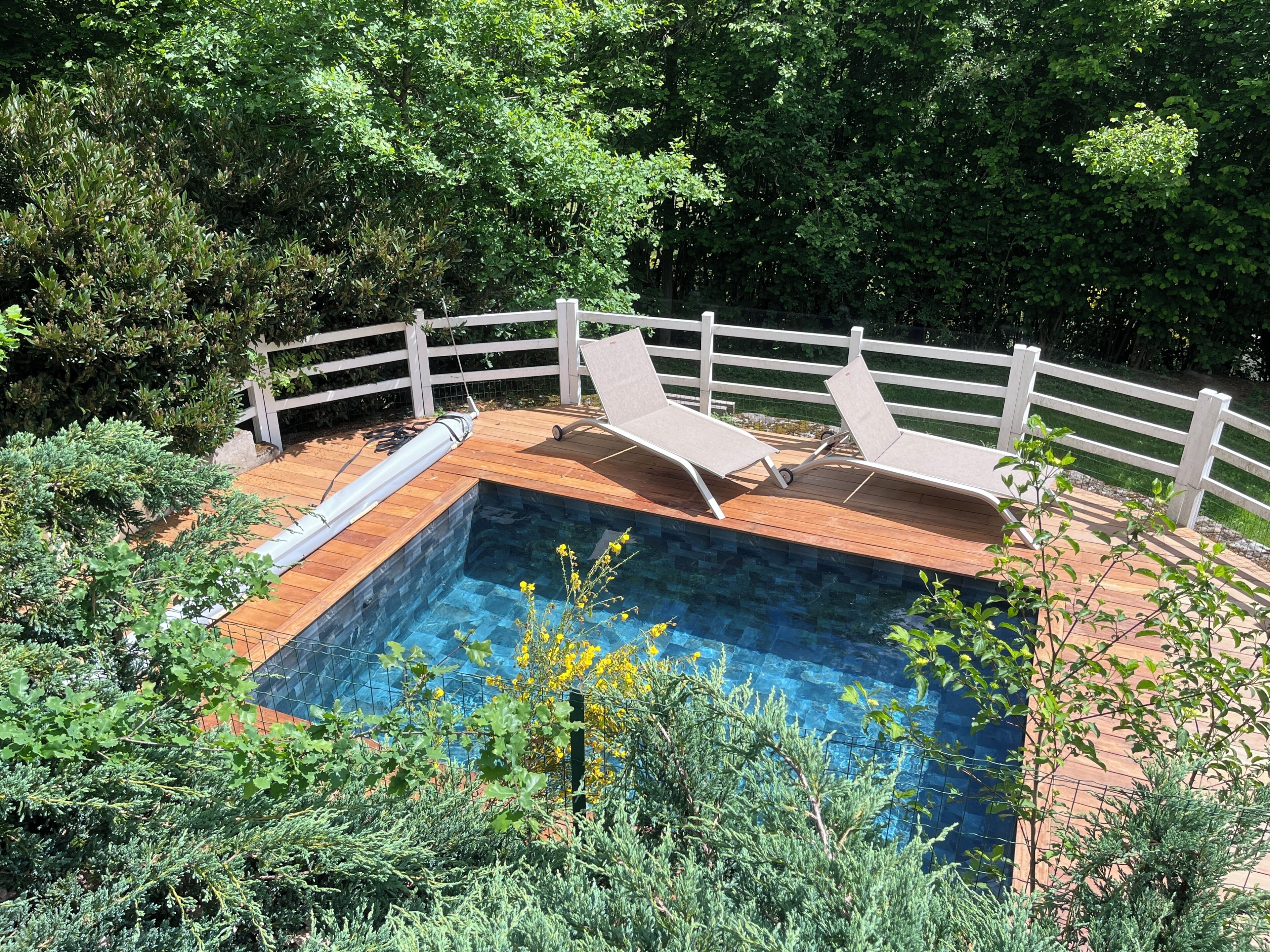 A swimming pool with a wooden deck, surrounded by a white fence, and lush greenery in the background.