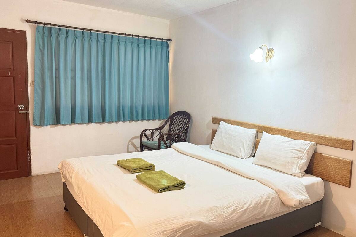 Una cama bien hecha con sábanas blancas y dos toallas verdes, un cabecero de madera, una silla y una ventana con cortinas azules.