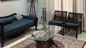 Un salon avec un canapé bleu, deux fauteuils en bois, une table basse en verre, un tableau accroché au mur, un ventilateur de plafond et une fenêtre avec des rideaux.