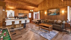 A cozy cabin interior with wooden walls, a kitchen area, a dining table, a sofa, and a foosball table. - Bernhardt Brown Bungalow #2583 (Big Bear City)