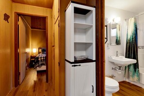 A wooden hallway leading to a bedroom with a bed, a white cabinet, and a bathroom with a sink and toilet. - Bernhardt Brown Bungalow #2583 (Big Bear City)