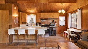 A wooden cabin interior with a kitchen island, bar stools, a dining table, and a sofa. - Bernhardt Brown Bungalow #2583 (Big Bear City)