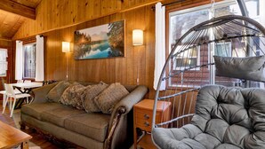 A wooden cabin interior with a sofa, a cushioned chair, a dining table, and a painting of a lake. - Bernhardt Brown Bungalow #2583 (Big Bear City)