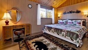 A bedroom with a wooden ceiling, a bed with a patterned quilt, a dresser, a mirror, a clock, and a window with curtains. - Bernhardt Brown Bungalow #2583 (Big Bear City)