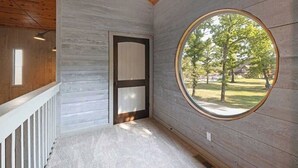 Une pièce avec un plafond en bois, une porte blanche et une grande fenêtre ronde donnant sur des arbres et une pelouse.