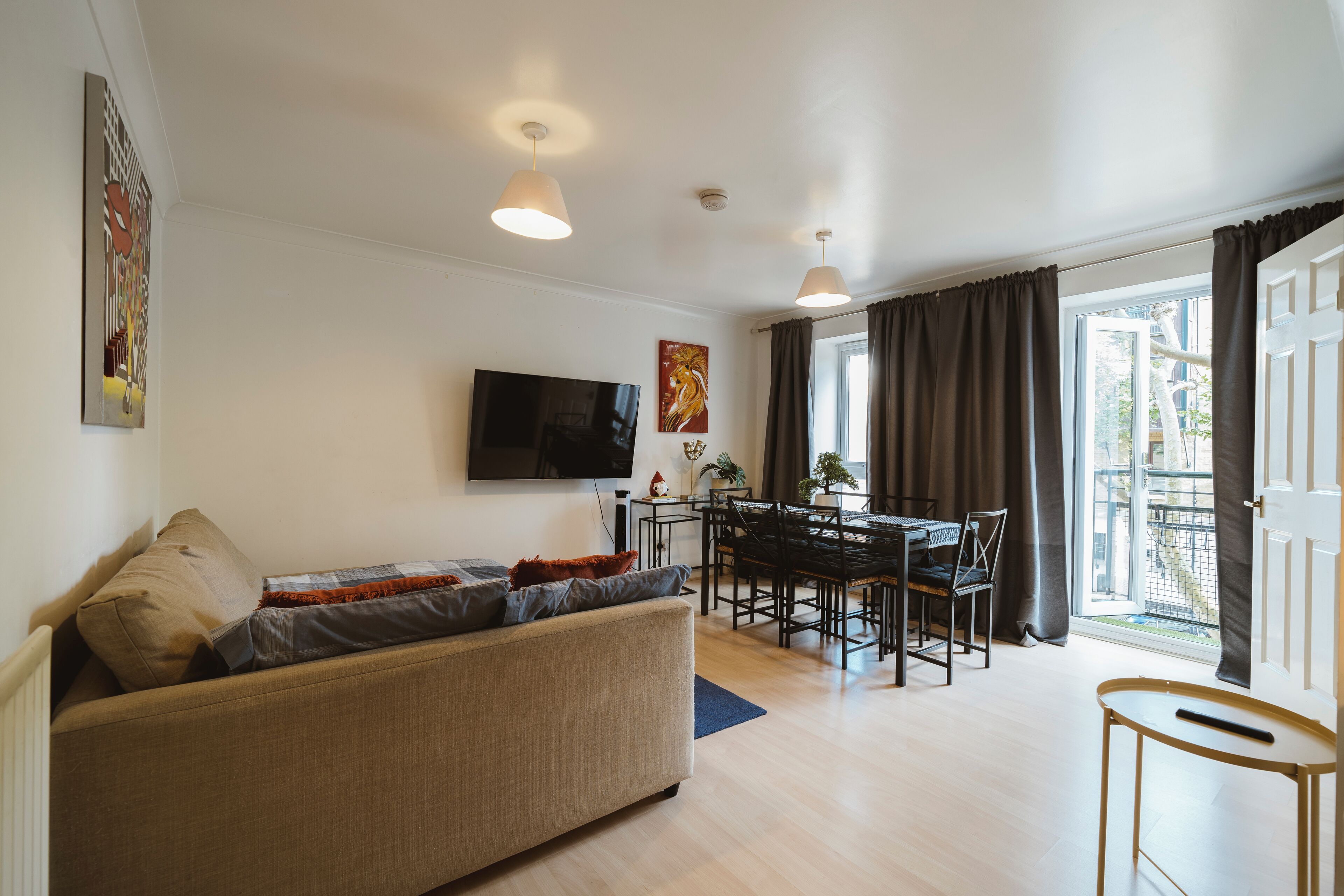 Ein modernes Wohnzimmer mit einer Couch, einem Esstisch, Stühlen, einem an der Wand befestigten Flachbildfernseher und einem Balkon mit Vorhängen.