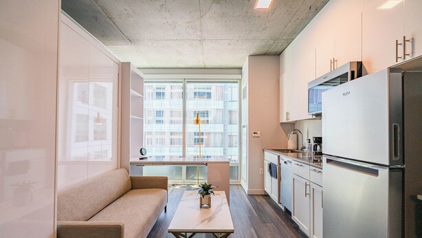 Een modern appartement met een zithoek, inclusief een bank en salontafel, een keuken met roestvrijstalen apparatuur en grote ramen met uitzicht op de skyline.