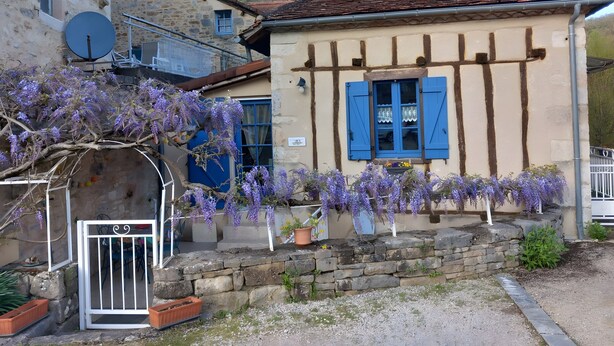 Ein traditionelles Fachwerkhäuschen mit einem steilen Satteldach, einem kleinen Fenster mit blauen Fensterläden und Glyzinien, die über den Eingang hängen.