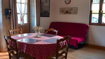 Un comedor con una mesa redonda, sillas, un sofá rojo, vigas de madera, una lámpara colgante y una ventana con cortinas.