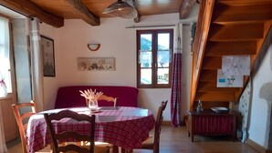 Ein Essbereich mit einem runden Tisch, Stühlen und einer roten Tischdecke. Es gibt ein Fenster mit Vorhängen, eine Holzbalkendecke und eine Treppe mit einem Holzaplauf.