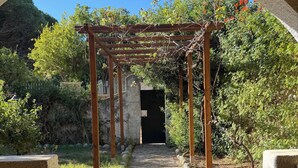 En stig leder fram till en mörk entré under en träpergola, omgiven av grönska och en klarblå himmel.