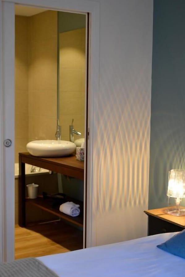 A hotel room with a bed, a bedside table with a lamp, a bathroom with a sink and mirror, and a wooden shelf with towels. - 5 chambres en ville (Clermont-Ferrand)