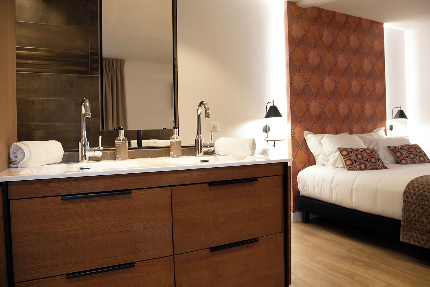 Ein Hotelzimmer mit einem großen Spiegel, einer Holzkommode mit Schubladen, einem Bett mit gemusterten Kissen und einem Nachttisch mit Lampe.