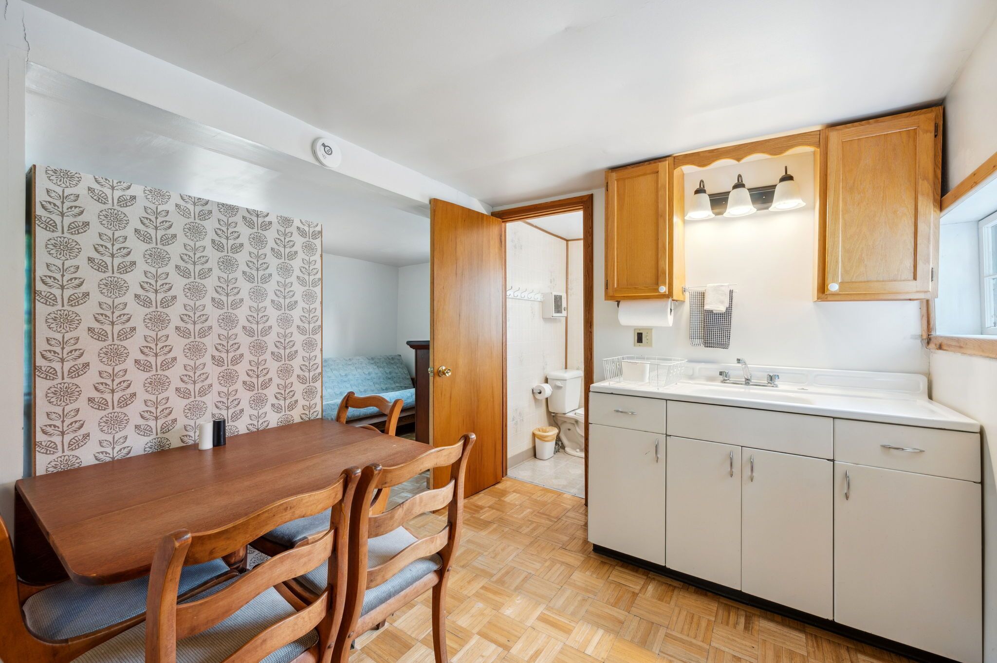 Een kleine keuken met houten kastjes, een spoelbak en een eethoek met een tafel en stoelen.