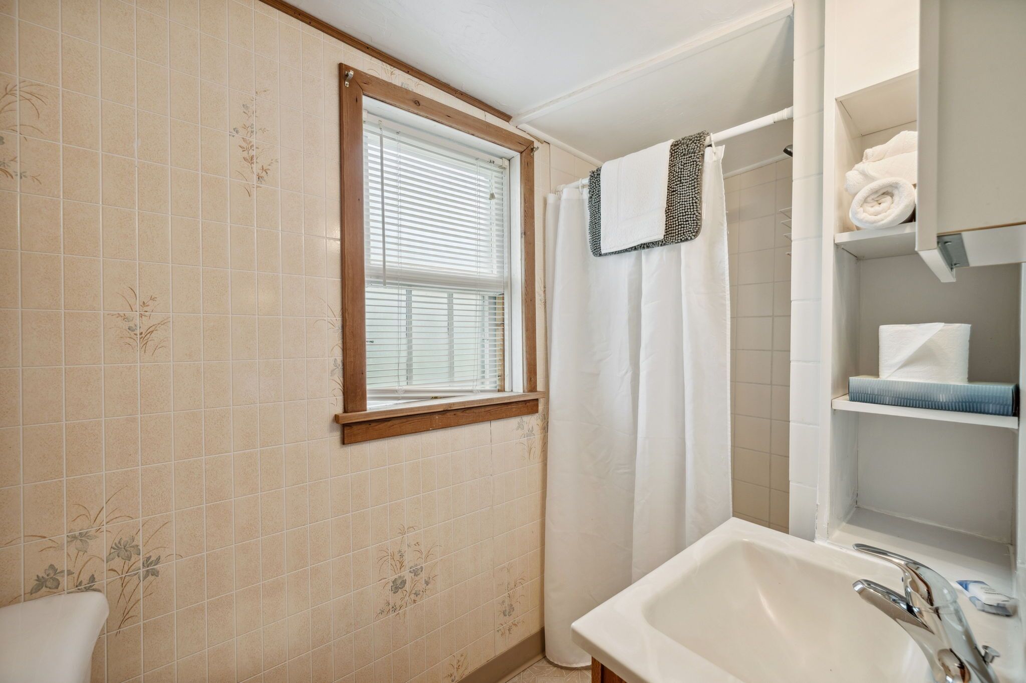 A bathroom with a window, a shower, a sink, and shelves with towels.