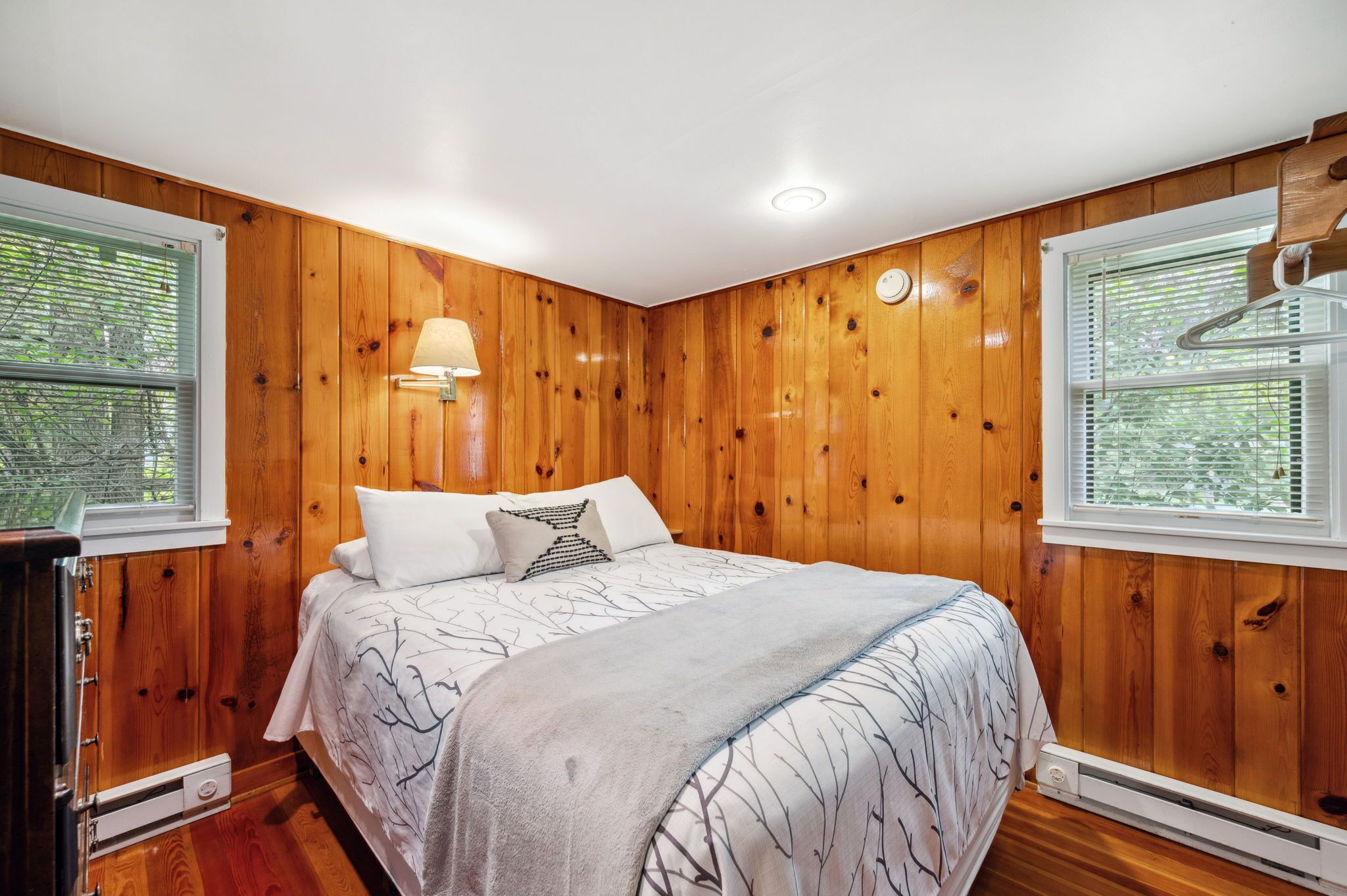 A bedroom with wooden walls, a bed with a patterned bedspread, a bedside lamp, and a heater under the bed.