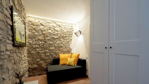 Una habitación con pared de piedra, un sofá negro con cojines amarillos, una puerta blanca y una lámpara fijada a la pared.