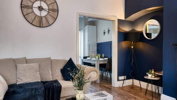 Ett vardagsrum med en soffa, ett soffbord och en väggklocka. Längre bak i rummet finns en matplats med ett bord och stolar.