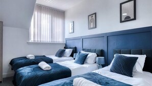 Ett hotellrum med tre sängar, alla med blått sänggavel, vita sängkläder och en hoprullad handduk. På väggen hänger två inramade konstverk, och det finns ett fönster med persienner.