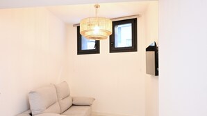 Een kleine, goed verlichte kamer met een beige bank, twee ramen met zwarte kozijnen en een aan de muur bevestigde televisie.