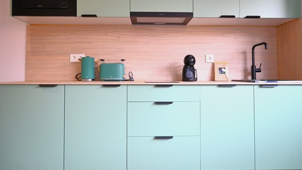 Een keuken met lichtgroene kastjes, een zwarte magnetron, een broodrooster, een waterkoker en een koffiezetapparaat.
